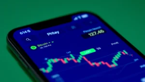 Close-up of a smartphone displaying a cryptocurrency exchange app with Bitcoin price charts and PHP conversion rates visible on the screen, showing real-time market data with candlestick patterns, modern minimalist design with blue and green color scheme, professional financial technology aesthetic, shallow depth of field